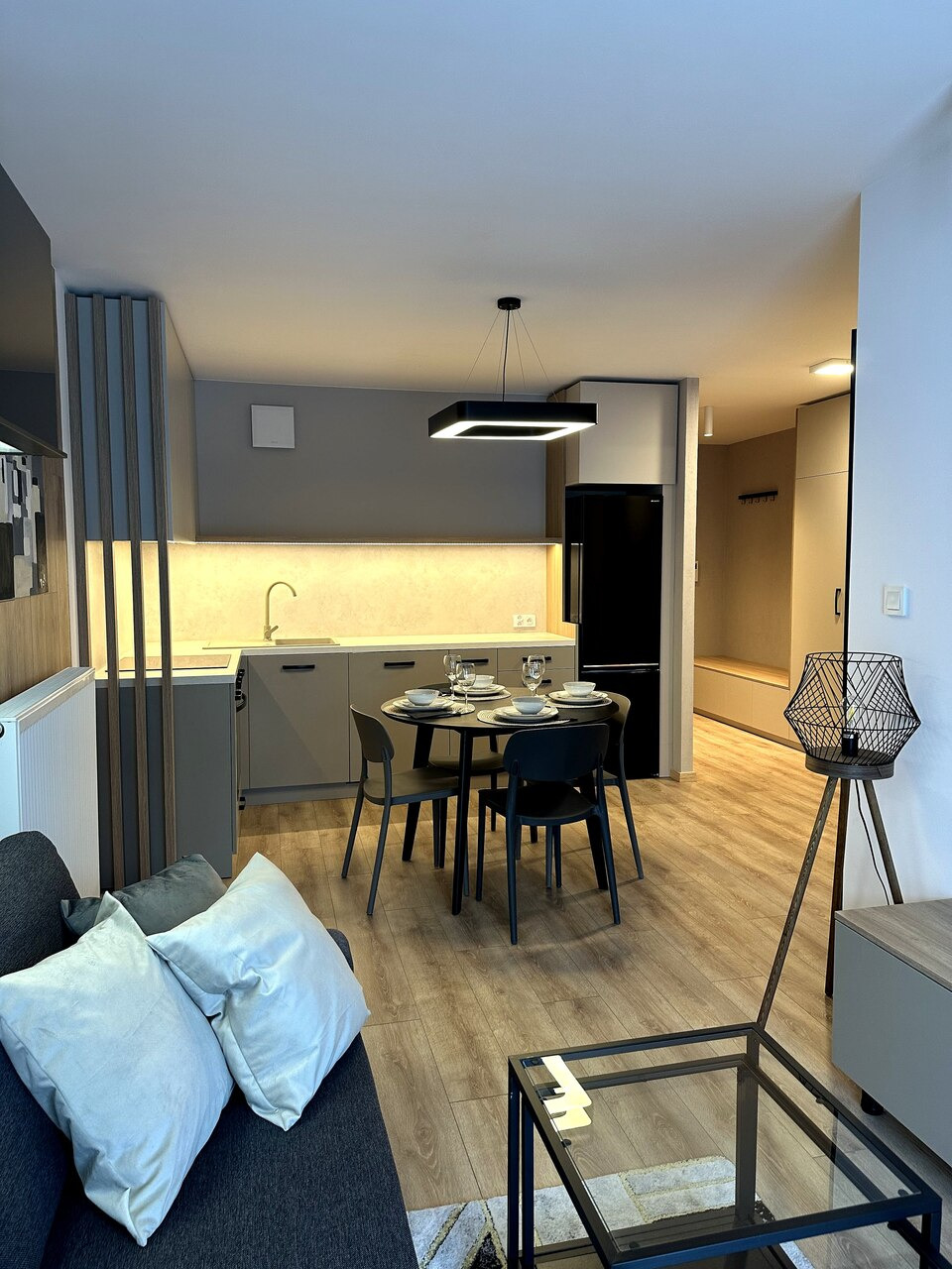 Nowoczesne wnętrze mieszkania z szarą zabudową kuchenną, okrągłym stołem z zastawą i czarną lodówką, widok z salonu.