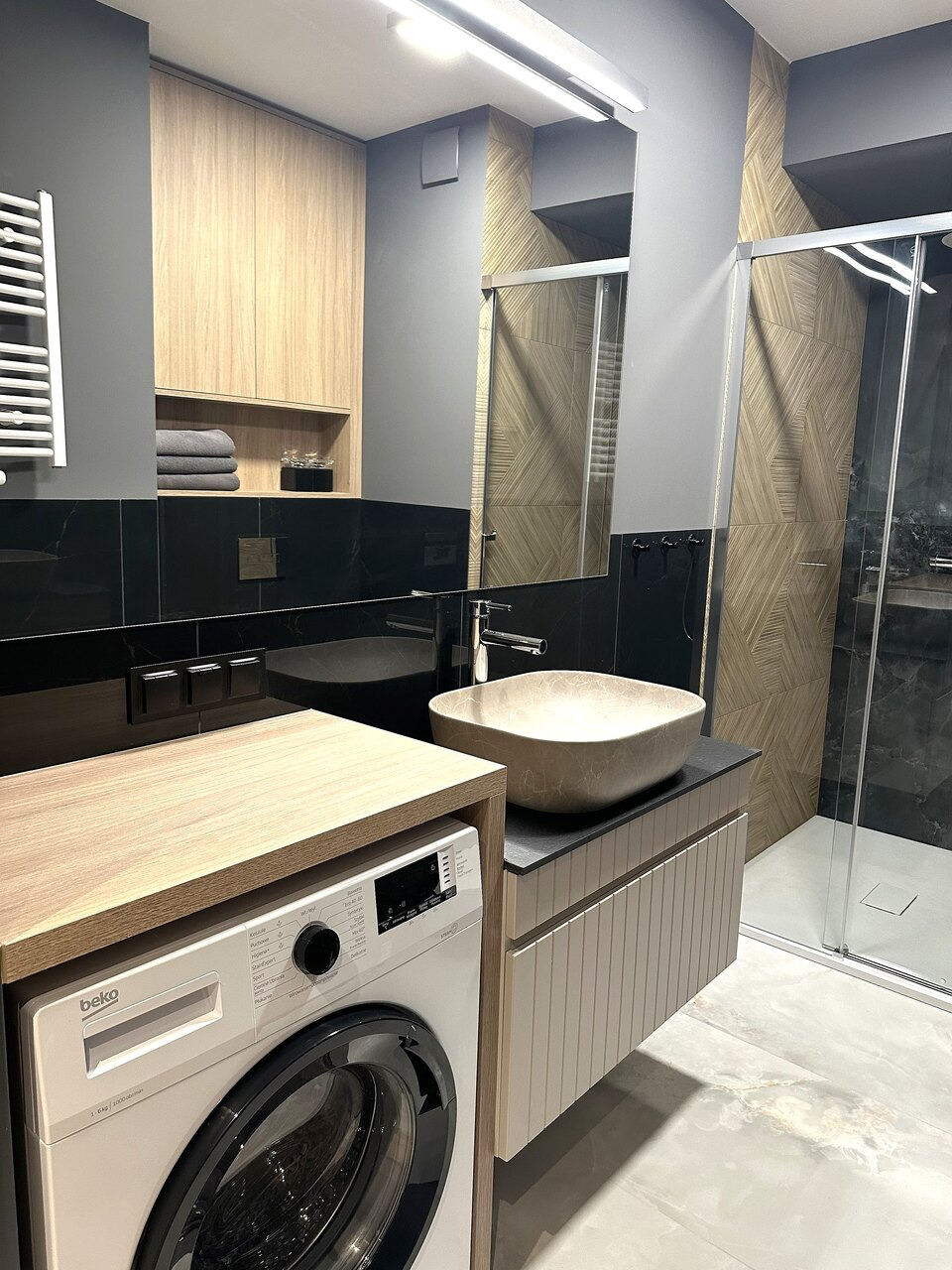 Nowoczesna łazienka w odcieniach szarości i drewna z pralką zabudowaną w szafce, umywalką nablatową w kształcie misy i kabiną prysznicową z geometrycznym wzorem na ścianach.