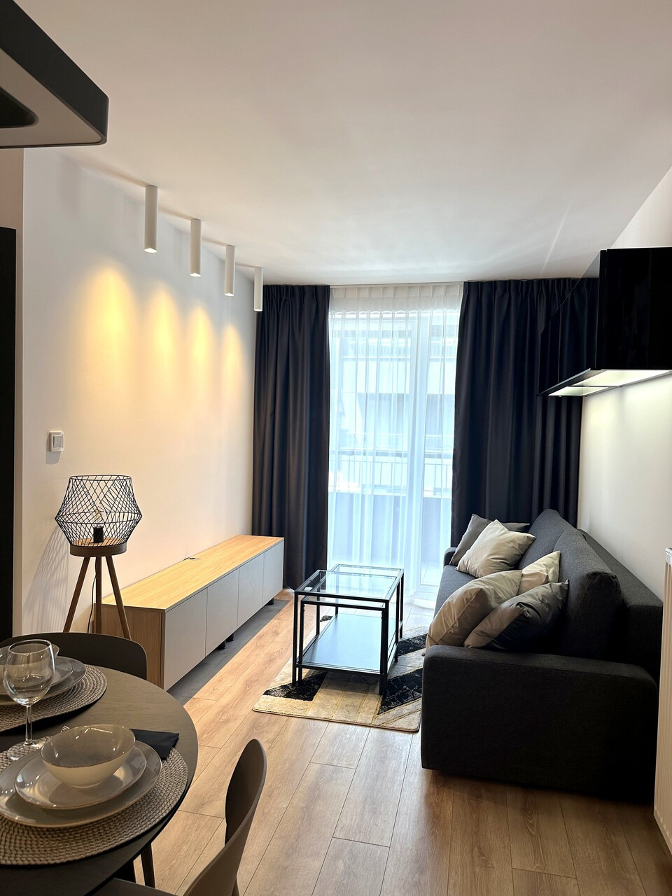 Nowoczesny salon z szarą sofą, stolikiem kawowym ze szklanym blatem, drewnianą szafką RTV i lampą na trójnogu, oświetlony listwą LED na ścianie.