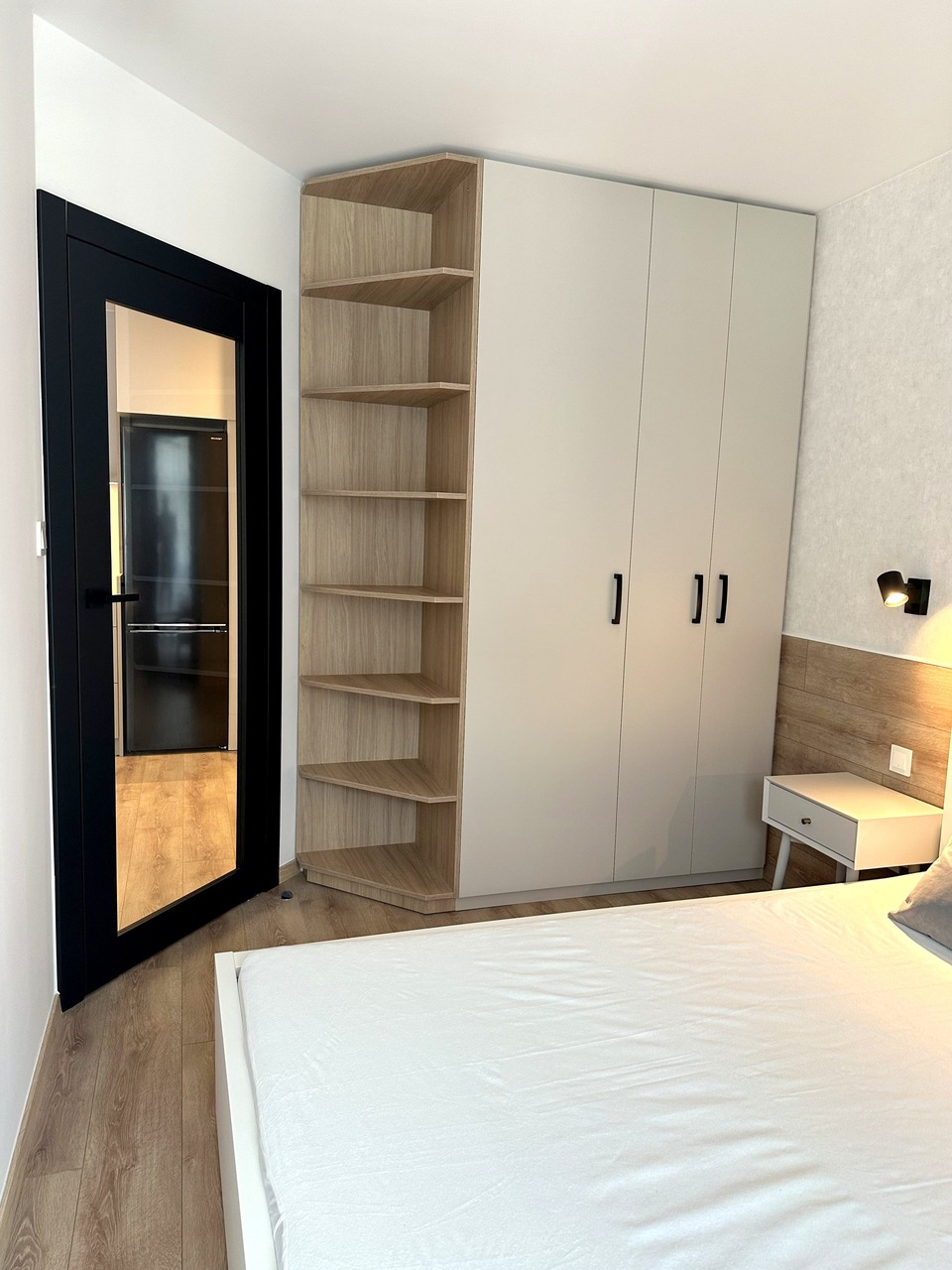 Jasna szafa z półkami narożnymi w sypialni, widoczny fragment łóżka z białą pościelą oraz czarne drzwi do innego pomieszczenia.