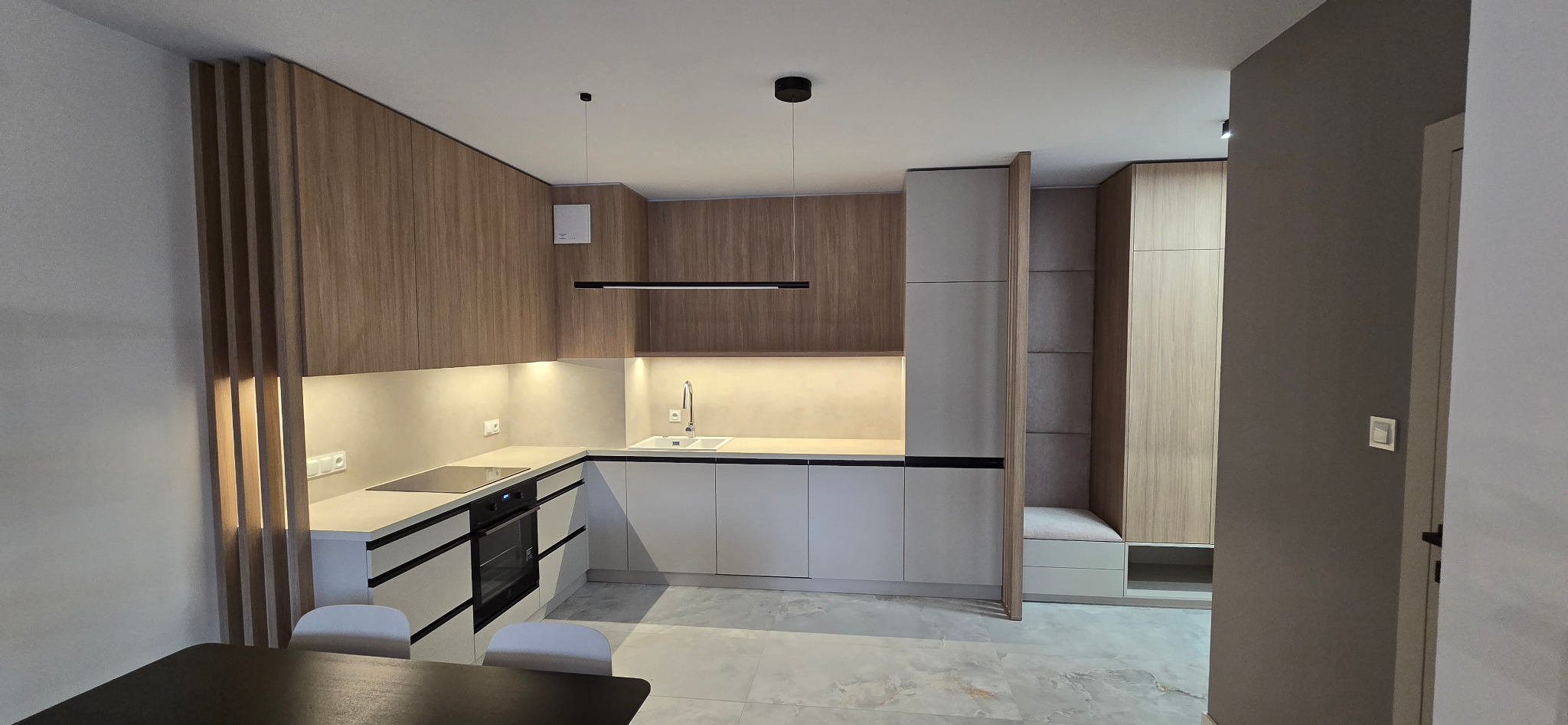 Nowoczesna kuchnia na wymiar z drewnianymi frontami górnych szafek i białymi dolnymi, czarne, minimalistyczne uchwyty, oświetlenie LED pod szafkami, widoczna część jadalni z czarnym stołem...