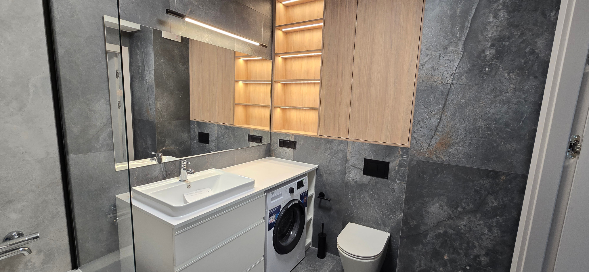 Nowoczesna łazienka z szarymi płytkami imitującymi kamień, białą umywalką nablatową, szafką z pralką w zabudowie, podświetlanymi półkami i dużym lustrem.