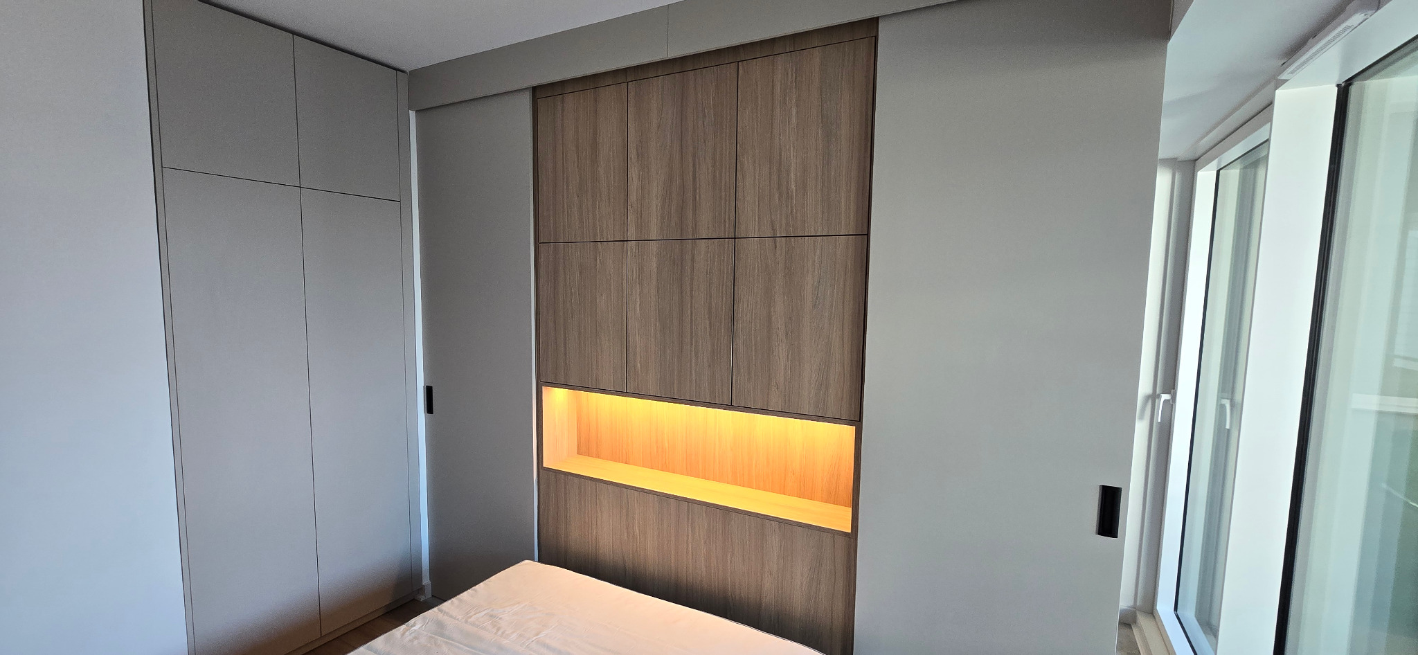 Nowoczesna szafa z drewnianymi frontami i wbudowanym oświetleniem LED, przylegająca do ściany z oknem balkonowym, widok z perspektywy łóżka.