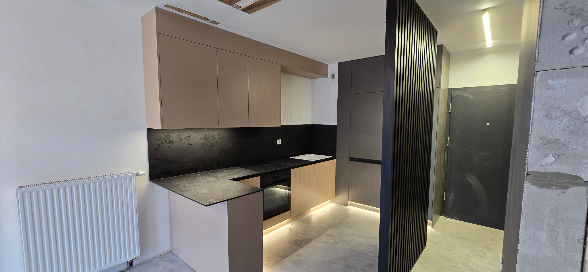 Nowoczesna kuchnia z beżowymi szafkami górnymi i dolnymi, czarnym blatem oraz ścianą nad blatem z efektem kamienia, oświetlona listwą LED pod szafkami, z elementami wykończenia w postaci pionowych...