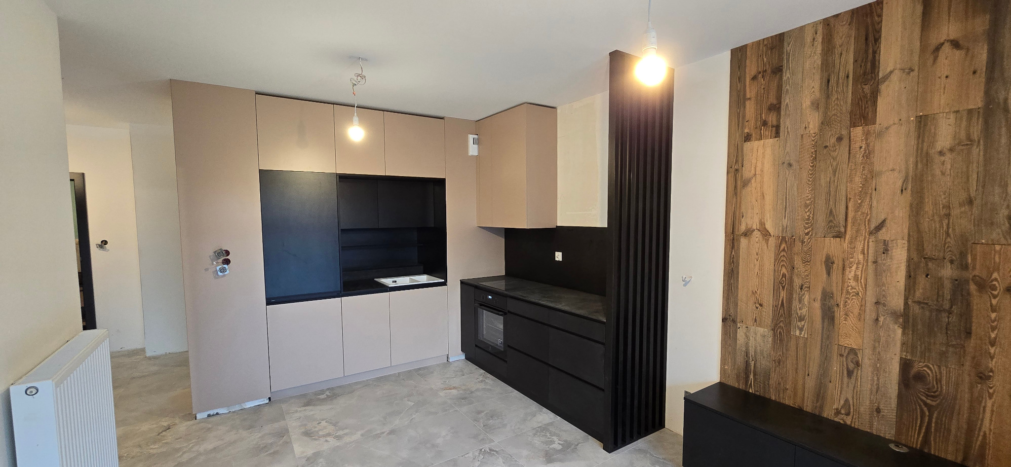 Nowoczesna kuchnia z beżowymi górnymi szafkami i czarnymi dolnymi, drewniana ściana w tle, surowy stan wykończenia podłogi.