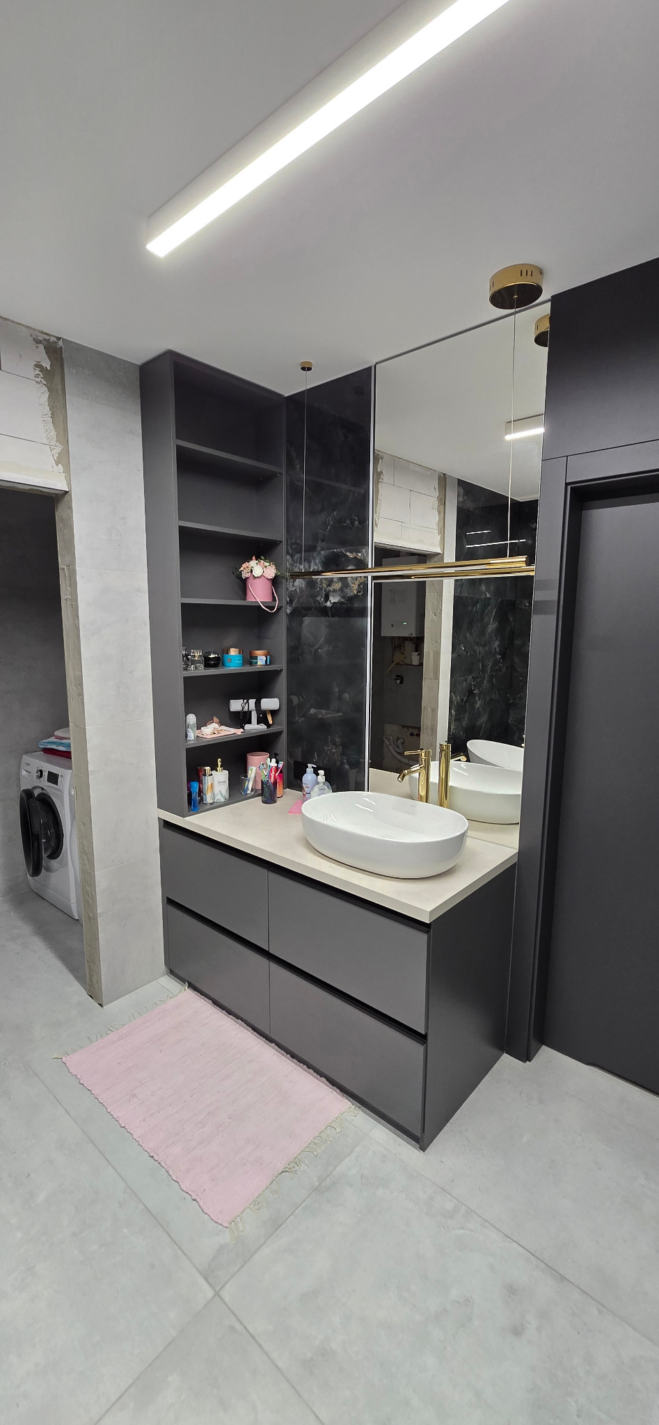 Szara zabudowa łazienkowa z szafkami i otwartym regałem, biała umywalka nablatowa, złota bateria, duże lustro, pralka w tle.