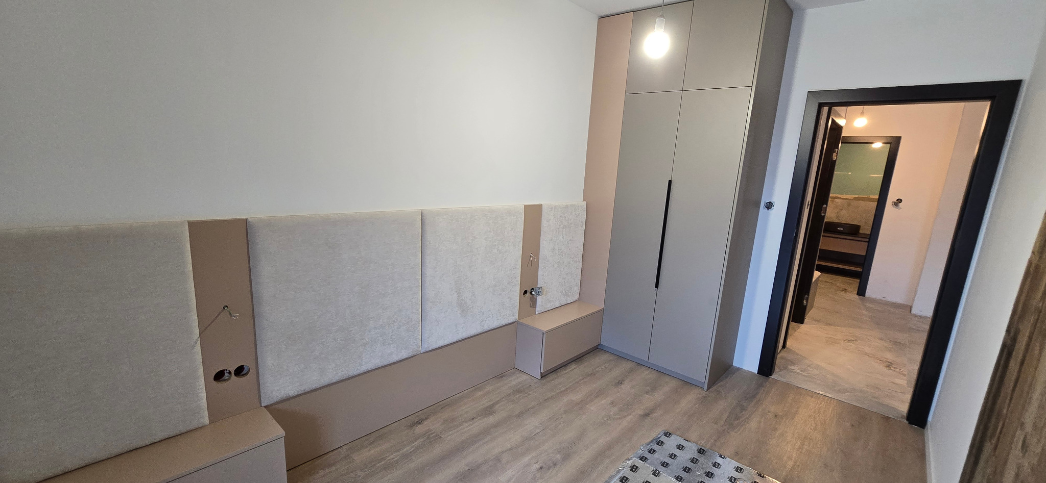 Nowoczesna sypialnia z zabudową: szara szafa na wymiar z długimi, czarnymi uchwytami, tapicerowany panel ścienny z beżowymi akcentami i wbudowanymi gniazdkami, widok na korytarz przez otwarte drzwi...