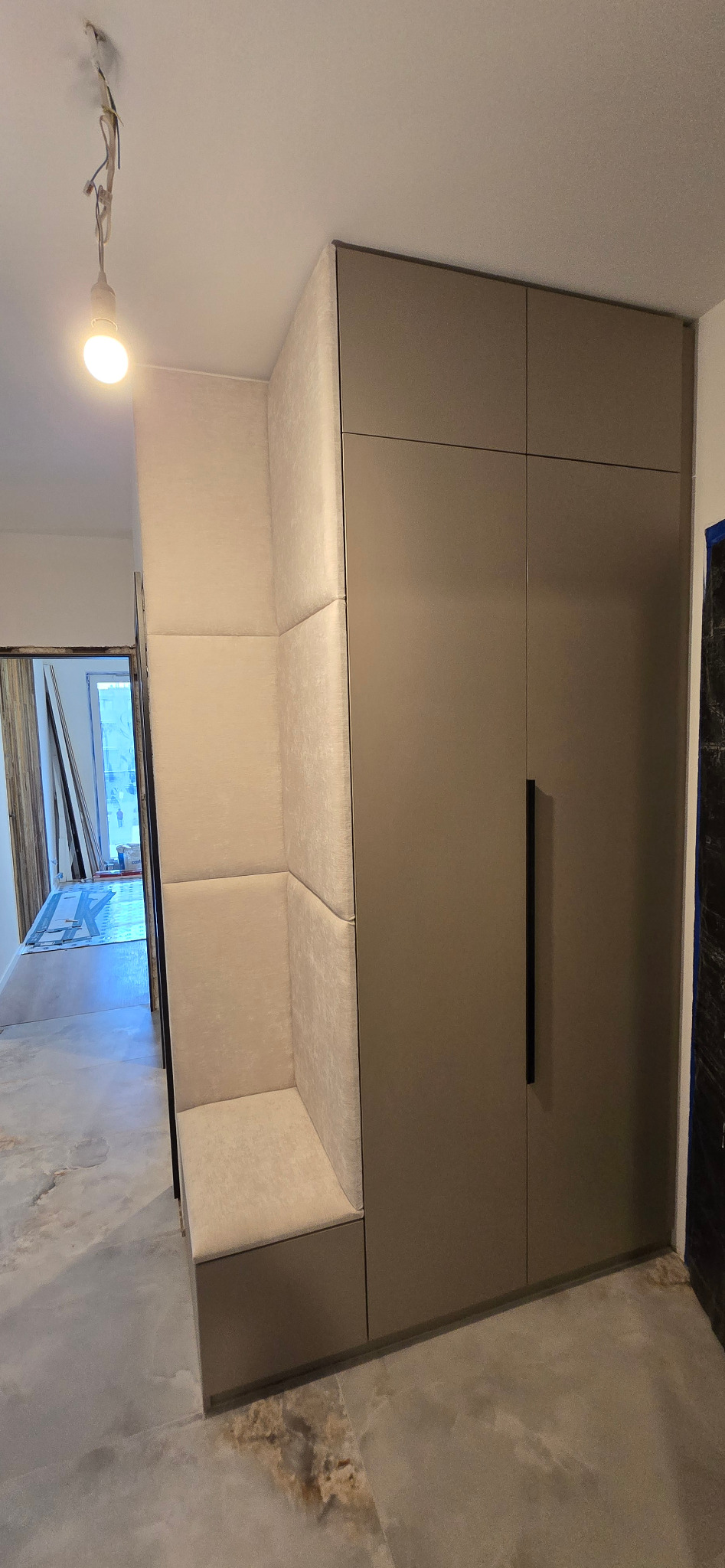 Wnęka z siedziskiem obok wysokiej szafy w neutralnych kolorach, widoczne niedokończone wykończenie podłogi i wiszące kable od oświetlenia sufitowego.