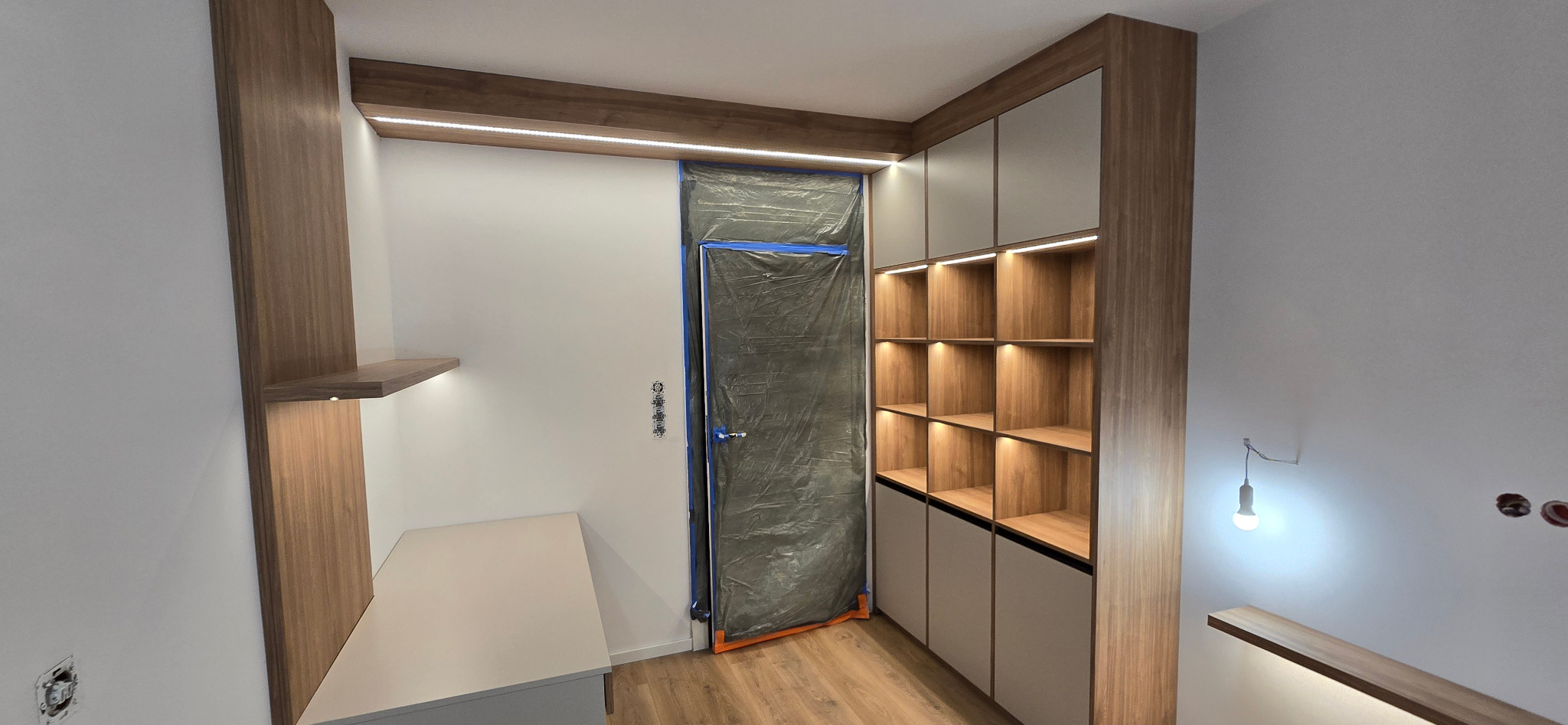 Wnętrze z zabudową meblową w trakcie prac wykończeniowych; widoczne regały z oświetleniem LED, biurko, zabezpieczone folią drzwi i wystające kable elektryczne.
