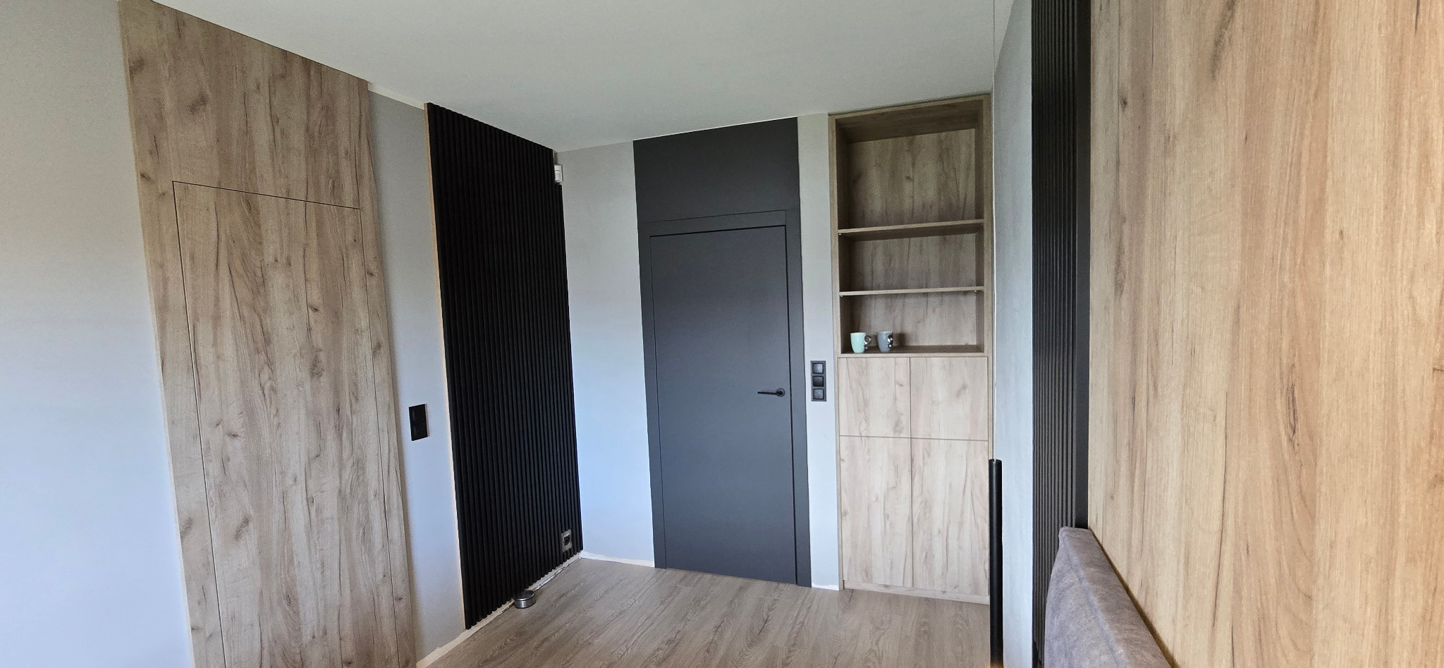 Wnętrze pokoju z drewnianą zabudową: szafa z ukrytymi drzwiami, czarny panel dekoracyjny, szara ściana z drzwiami i regał z szafką w kolorze drewna.