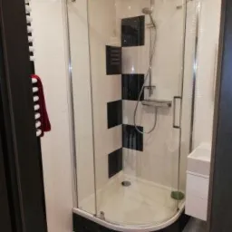 Kabina prysznicowa narożna z czarno-białymi płytkami w szachownicę na ścianie, widok z zewnątrz, z widocznym fragmentem czerwonego dywanika i ręcznika.