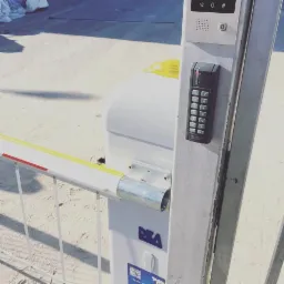 Szlaban automatyczny z panelem kodowym i domofonem na słupku, widoczne logo producenta DEA, oświetlenie słoneczne z cieniem.