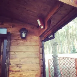 Drewniana elewacja domu z zamontowanym wideodomofonem w obudowie oraz lampą w stylu retro, widok na las w tle.