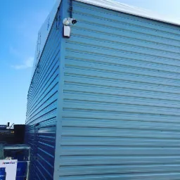 Zewnętrzna kamera monitoringu zamontowana na bocznej, blaszanej ścianie budynku Amerigas, widok z dołu w słoneczny dzień.