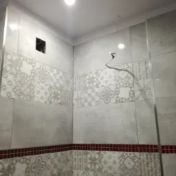 Ściana łazienki w Ciechanowie wyłożona szarymi płytkami z dekoracyjnym wzorem, widoczna czerwona bordiura, otwór w ścianie i wystające przewody elektryczne.