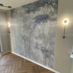 Remar - Pokój po remoncie: ściana z fototapetą imitującą las we mgle, sąsiadująca z panelem ściennym z pionowymi paskami, oświetlona nowoczesnymi lampami ściennymi, parkiet ułożony w jodełkę.