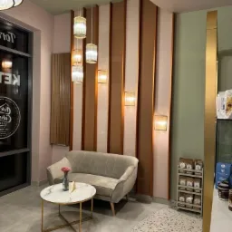 Nowoczesne wnętrze kawiarni z beżową sofą, okrągłym stolikiem z marmurowym blatem i złotą podstawą, wiszącymi lampami z abażurami w kształcie cylindrów oraz panelami dekoracyjnymi na ścianie.