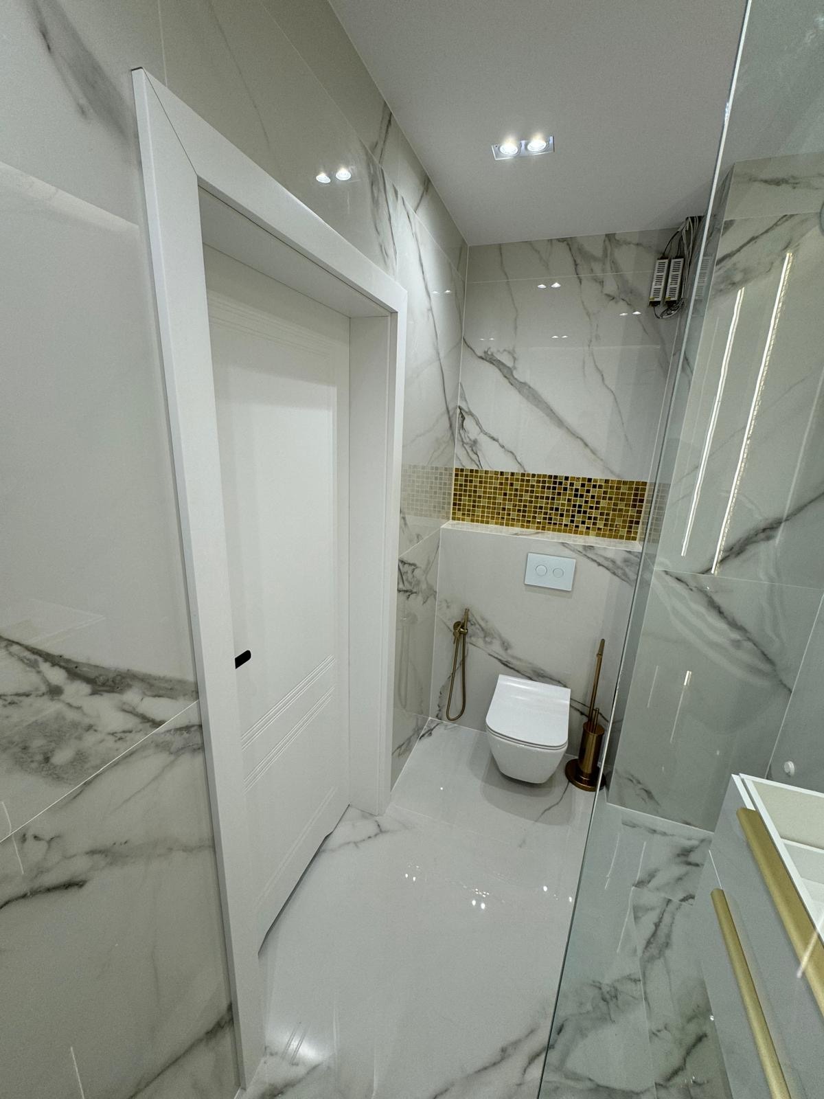 Wnętrze wykończonej łazienki z marmurowymi płytkami, złotą mozaiką, toaletą wiszącą i szklaną kabiną prysznicową, widok na białe drzwi z czarną klamką.