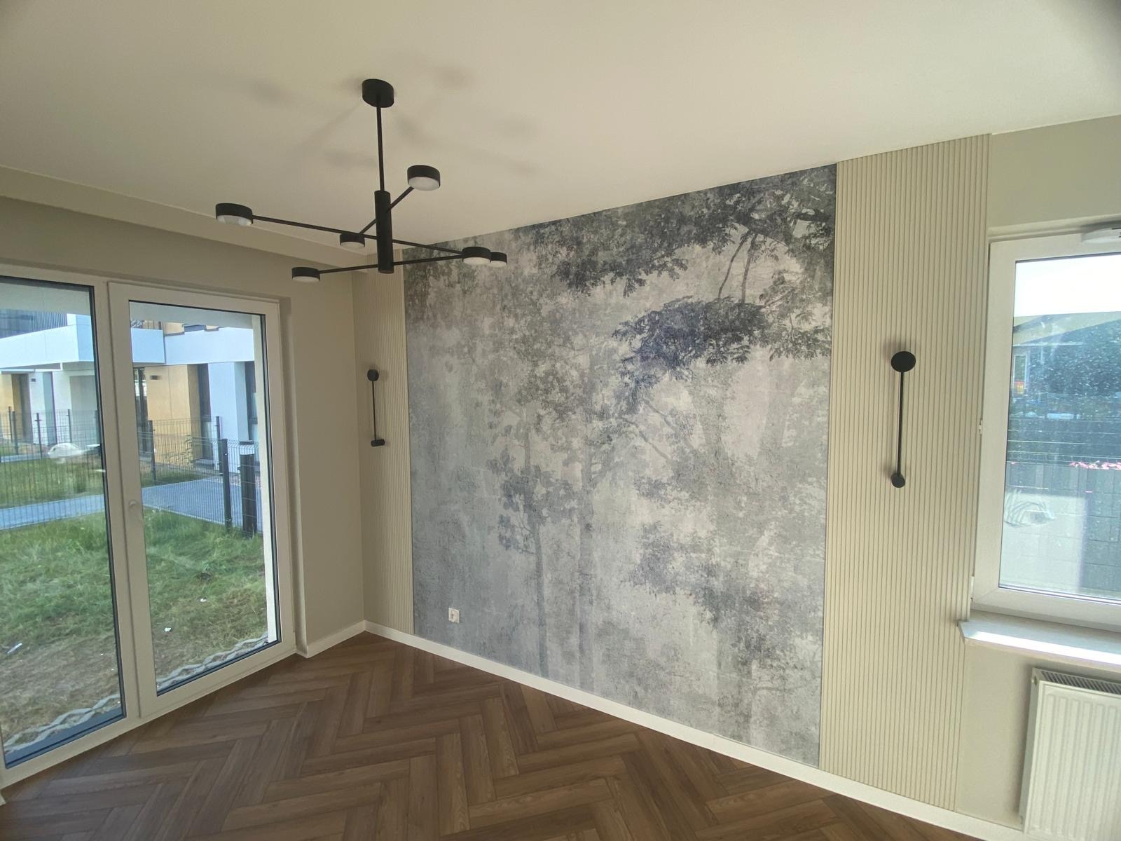 Pokój z parkietem w jodełkę, ścianą z fototapetą leśną i pionowymi panelami ściennymi, oświetlony nowoczesną lampą sufitową.