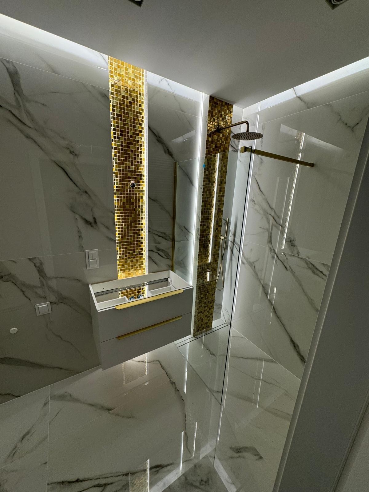 Nowoczesna łazienka z kabiną prysznicową typu walk-in, wykończona białymi płytkami imitującymi marmur z czarnymi żyłami, złotymi mozaikami i podświetleniem LED. Szafka pod umywalkę w kolorze szarym...