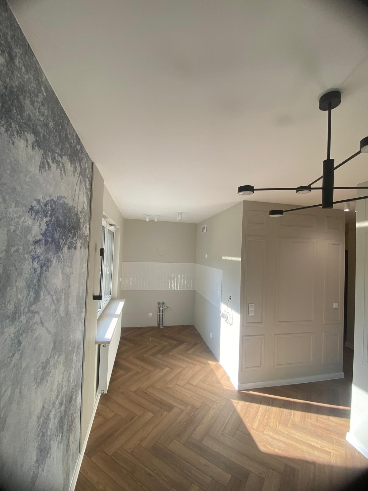 Nowoczesne wnętrze w trakcie wykańczania: parkiet ułożony w jodełkę, ściana z fototapetą imitującą las, białe płytki do połowy wysokości ściany, designerska lampa sufitowa, dekoracyjna ściana...