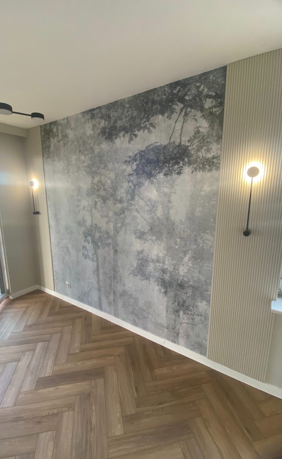Pokój po remoncie: ściana z fototapetą imitującą las we mgle, sąsiadująca z panelem ściennym z pionowymi paskami, oświetlona nowoczesnymi lampami ściennymi, parkiet ułożony w jodełkę.