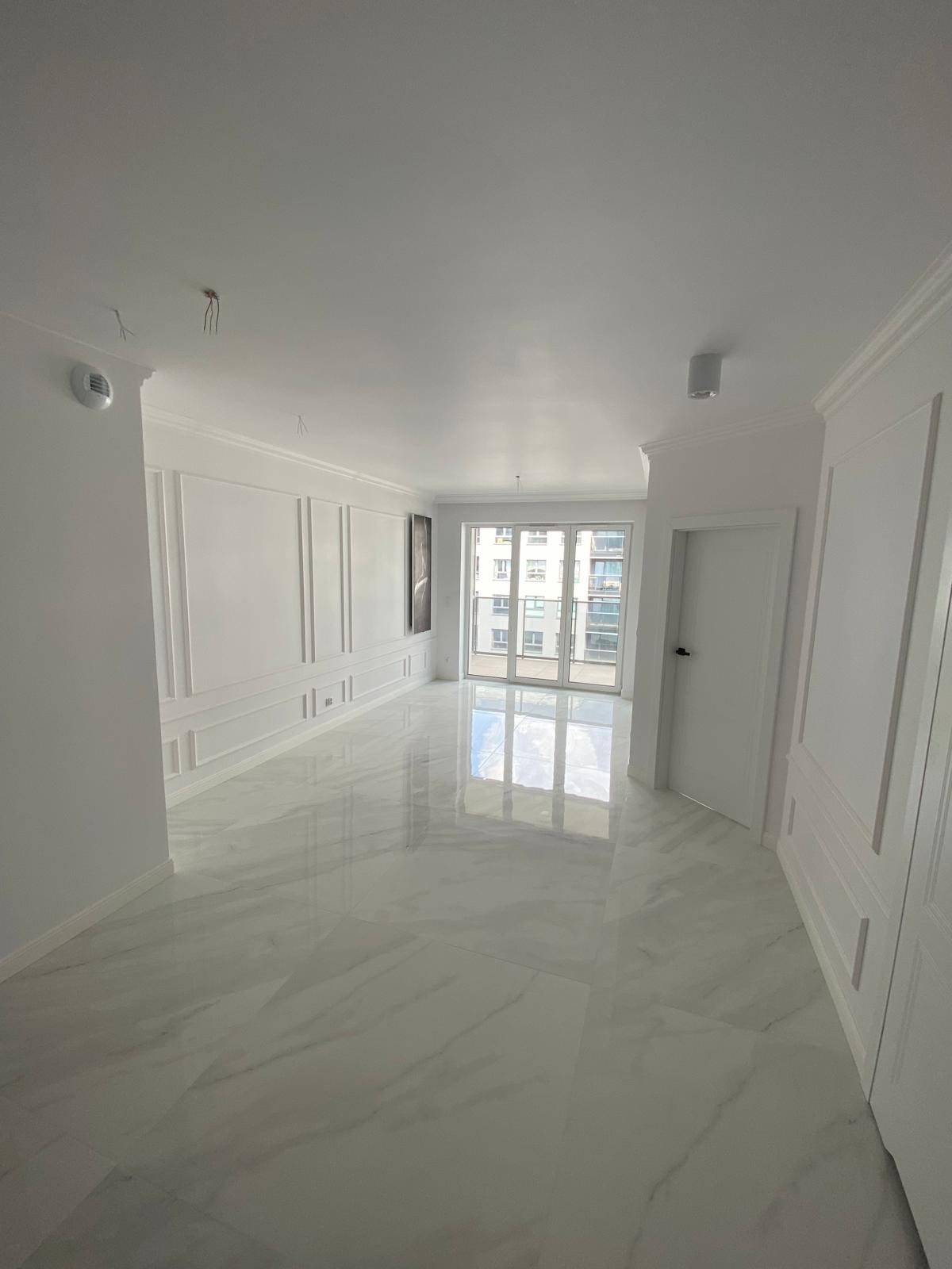 Jasne, przestronne wnętrze po remoncie z białymi ścianami zdobionymi sztukaterią, błyszczącą podłogą imitującą marmur i dużym oknem balkonowym; widać fragmenty wystających kabli na suficie.