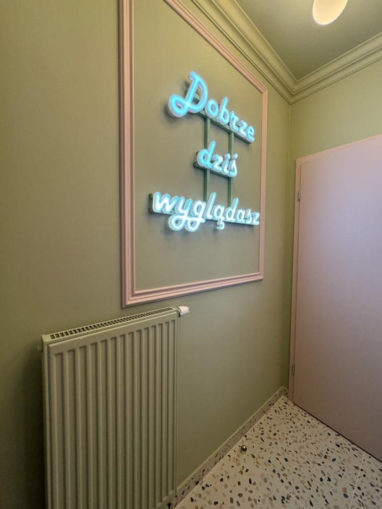 Ściana w odcieniach zieleni z grzejnikiem i neonowym napisem w ramce 'Dobrze dziś wyglądasz', podłoga w terazzo.