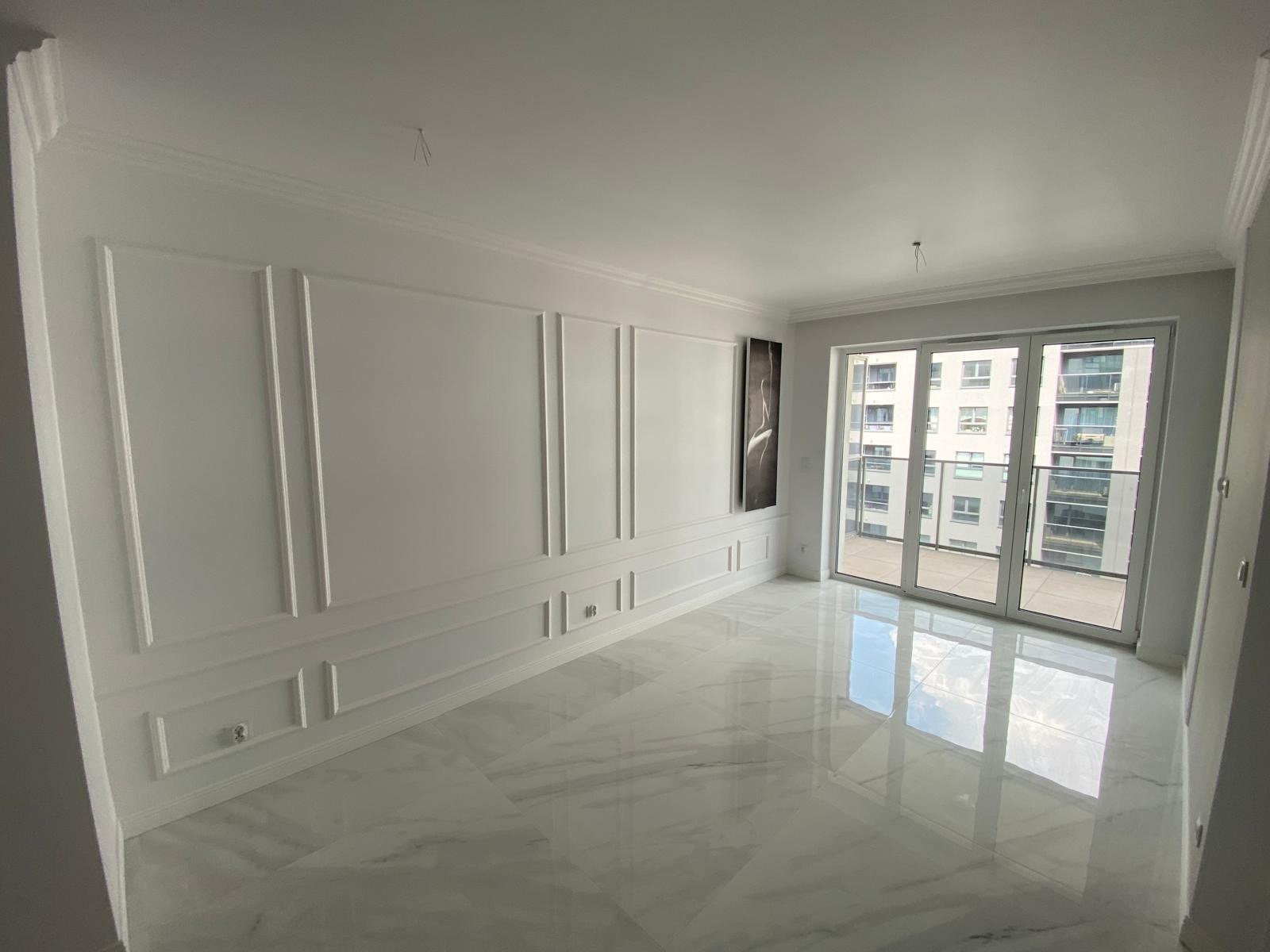 Jasne, puste pomieszczenie z białymi ścianami zdobionymi sztukaterią, marmurową podłogą odbijającą światło i balkonem z widokiem na budynek mieszkalny.