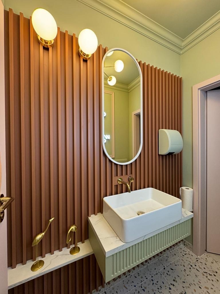 Nowoczesna łazienka z umywalką nablatową, złotą armaturą i dekoracyjnymi figurkami ptaków na marmurowym blacie, oświetlona przez okrągłe lampy ścienne i lustro z podświetleniem.