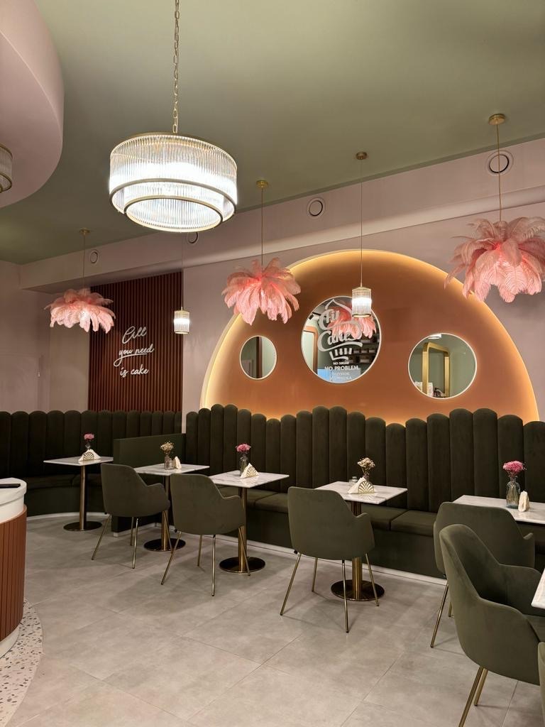 Wnętrze kawiarni w odcieniach pastelowych różów i zieleni, z dekoracyjnymi lampami z piórami, marmurowymi stolikami i pluszowymi siedziskami w stylu art deco.