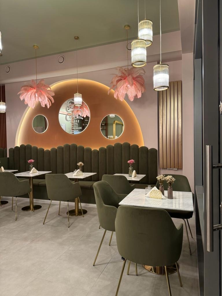 Eleganckie wnętrze kawiarni z zielonymi krzesłami i marmurowymi stolikami, oświetlone dekoracyjnymi lampami z różowymi piórami, z elementami wykończenia w odcieniach różu i złota.