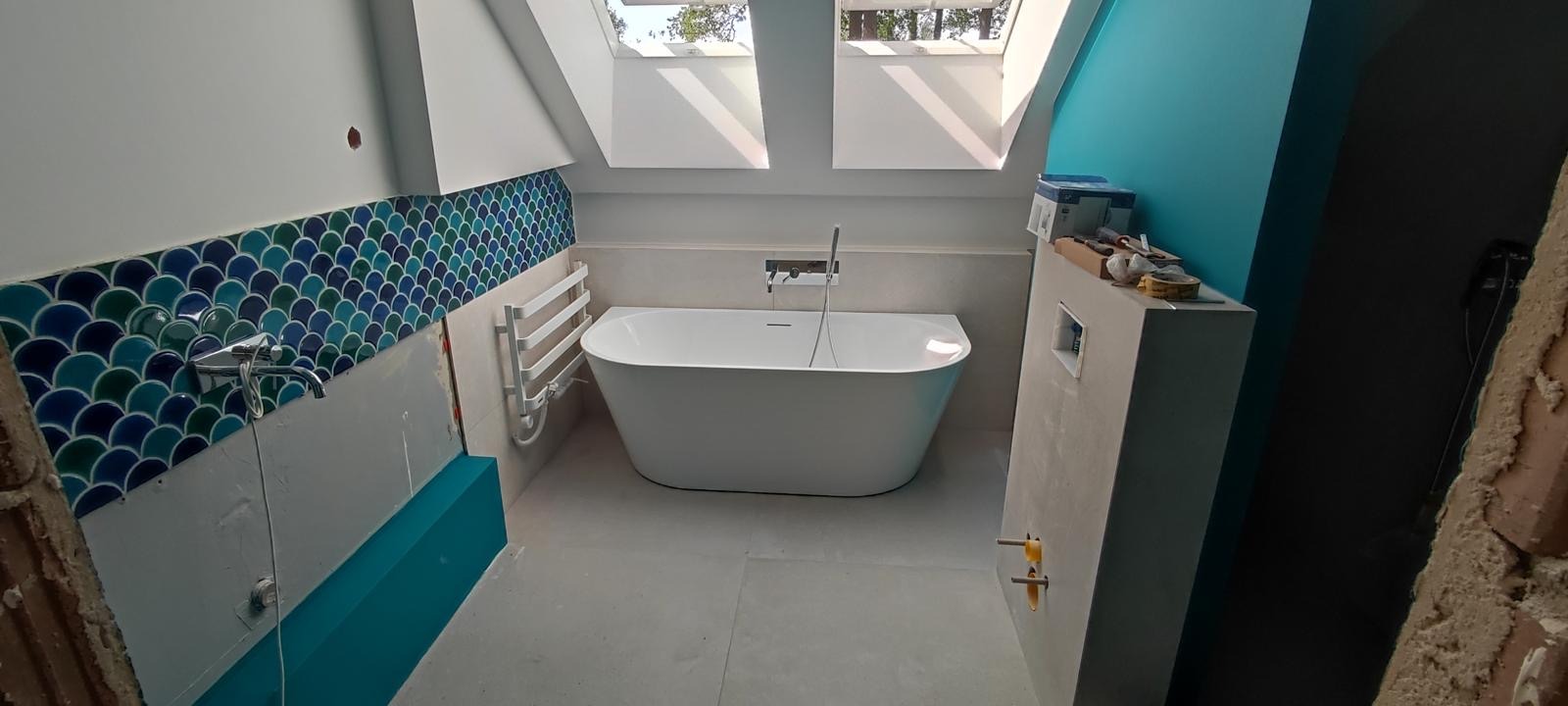 Łazienka na poddaszu w trakcie prac wykończeniowych: biała wolnostojąca wanna, grzejnik drabinkowy, fragment ściany z niebiesko-zieloną mozaiką w kształcie rybiej łuski, okna dachowe, surowy beton...