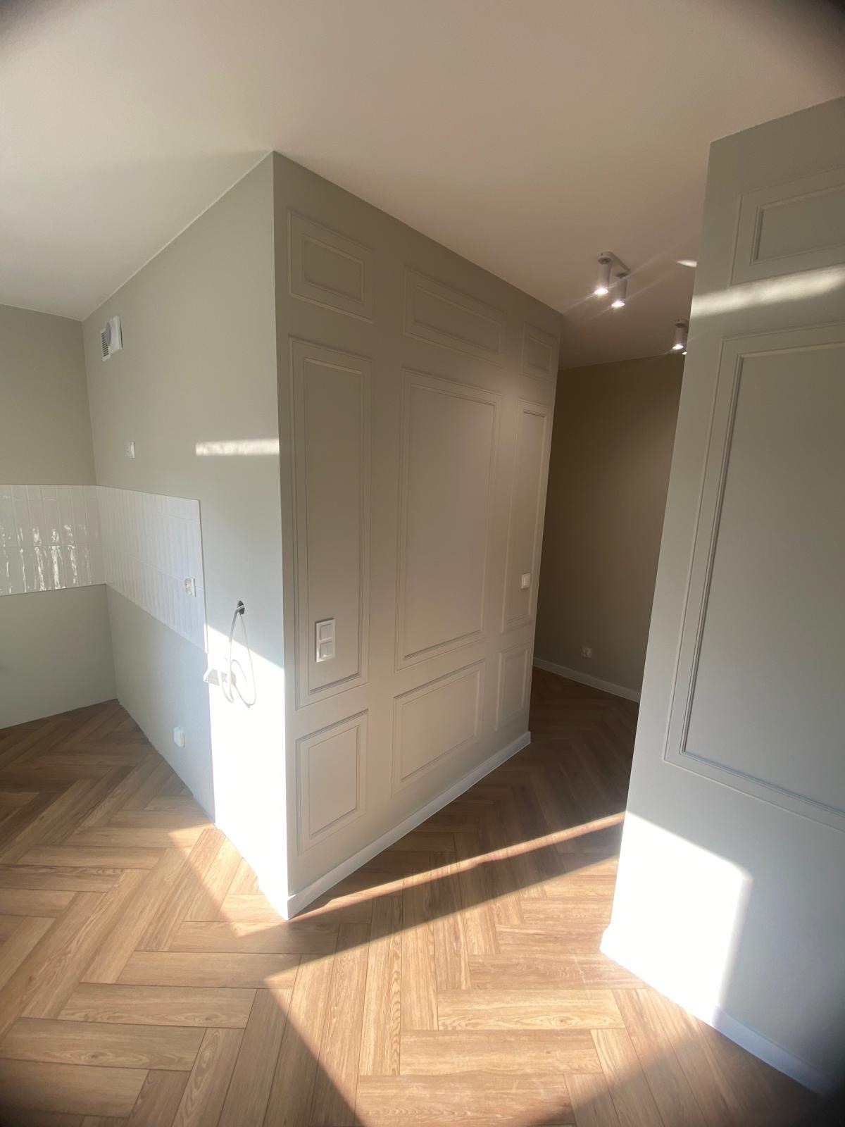 Jasne wnętrze po remoncie z panelami ułożonymi w jodełkę, dekoracyjnymi panelami ściennymi i białymi płytkami w kuchni, oświetlone promieniami słonecznymi.