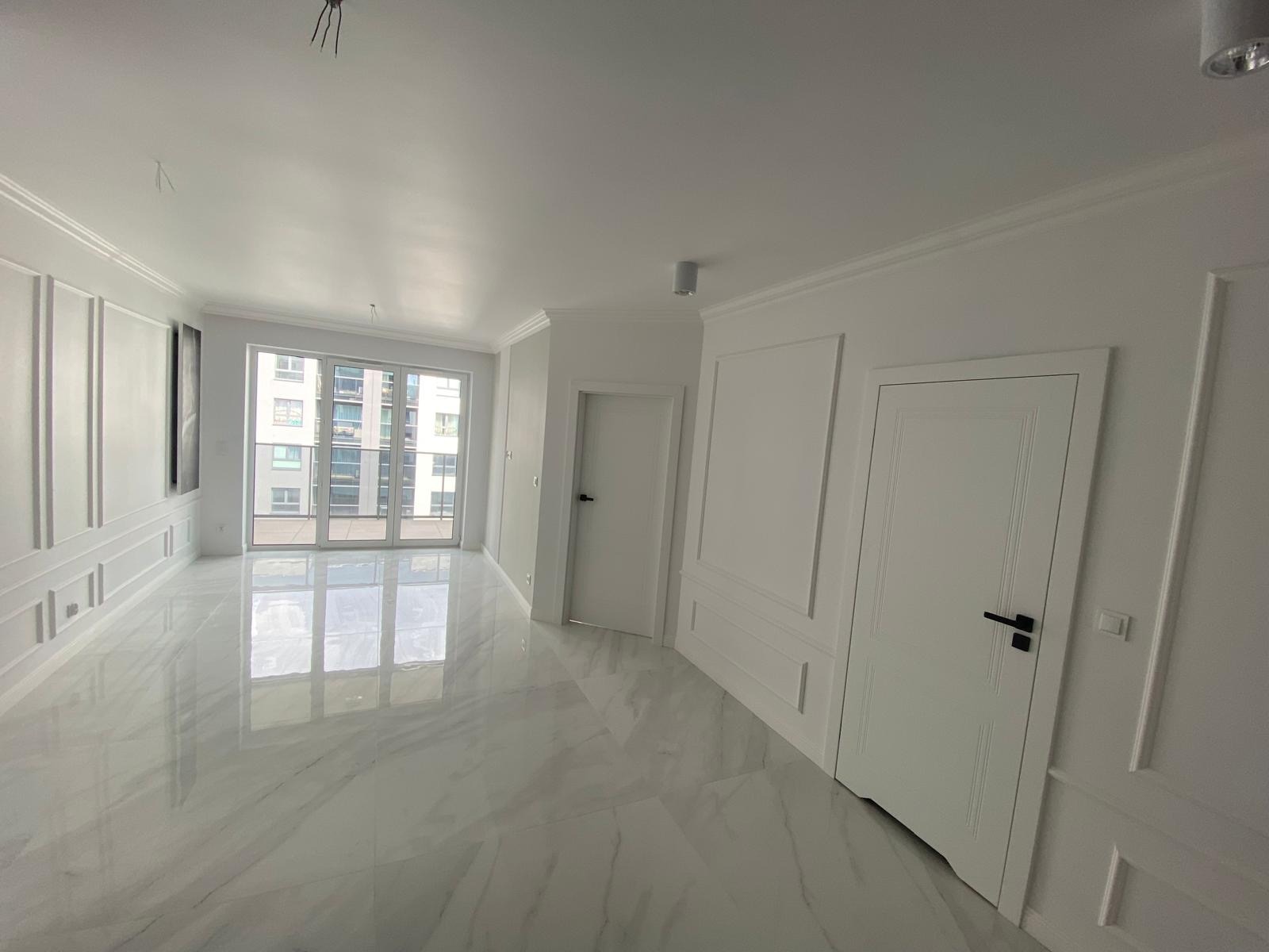 Jasne, przestronne wnętrze z marmurową podłogą, dekoracyjnymi panelami ściennymi i białymi drzwiami, widok na okno balkonowe z wysokim budynkiem w tle.