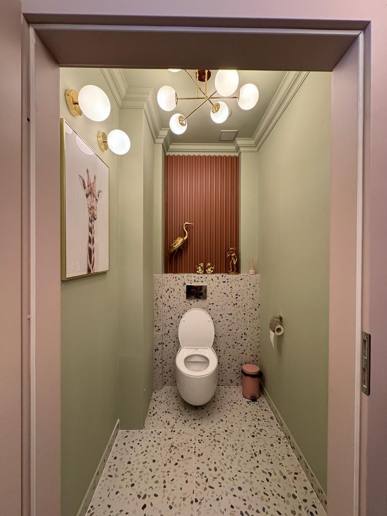 Nowoczesna łazienka z toaletą wiszącą, mozaikową ścianą i podłogą terrazzo, oświetlona lampą sufitową w stylu glamour i kinkietami, z dekoracjami w postaci złotych figurek ptaków i obrazu z żyrafą.