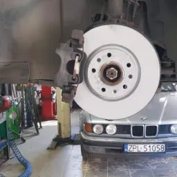 Nowa tarcza hamulcowa zamontowana w samochodzie BMW na podnośniku warsztatowym, widoczne elementy zawieszenia i zacisk hamulcowy, na podłodze leży stara tarcza i opona.