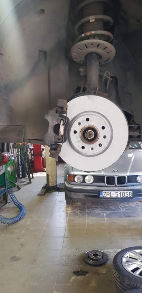 Nowa tarcza hamulcowa zamontowana w samochodzie BMW na podnośniku warsztatowym, widoczne elementy zawieszenia i zacisk hamulcowy, na podłodze leży stara tarcza i opona.