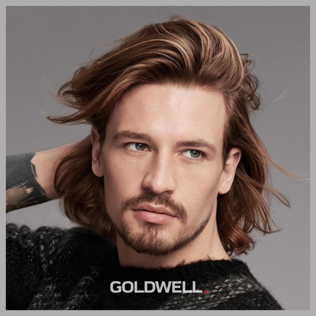 Mężczyzna z długimi, brązowymi włosami z blond pasemkami, stylizowanymi na bok, trzymający rękę za głową, widoczne tatuaże na ramieniu, prezentacja marki Goldwell.
