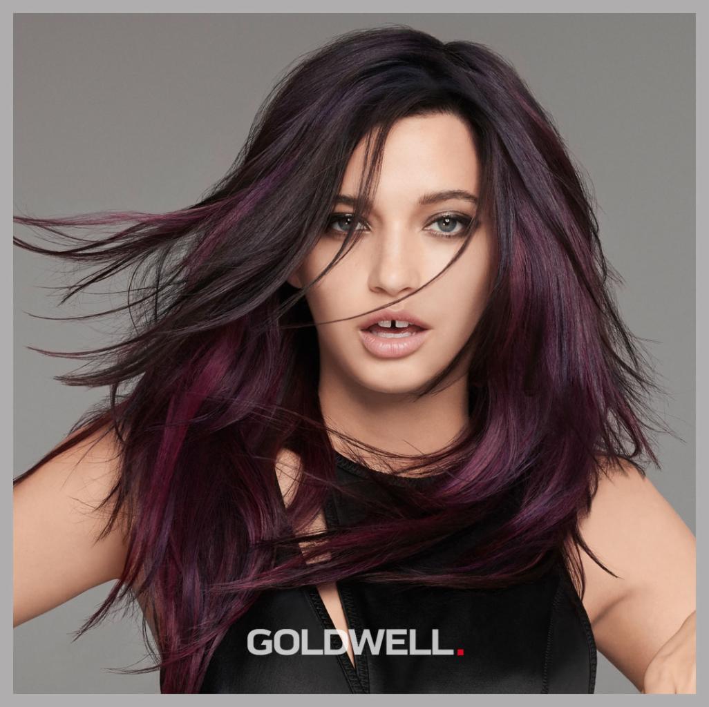 Modelka z ciemnymi włosami z purpurowymi pasemkami, włosy w lekkim ruchu, logo Goldwell na dole zdjęcia.