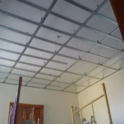 Montaż sufitu podwieszanego z płyt kartonowo-gipsowych na metalowej konstrukcji w pomieszczeniu z widoczną drabiną i ościeżnicą drzwi.