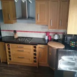 Jasna, drewniana zabudowa kuchenna z czarnym blatem, widoczny fragment płyty gazowej, zlewozmywak ze stali nierdzewnej, białe płytki z czerwonym paskiem oraz dekoracja ścienna w kształcie skutera.