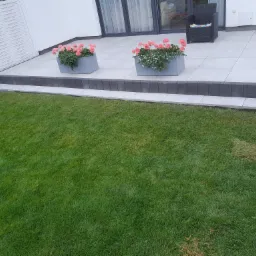 Nowoczesny taras z jasnych płyt, obrzeża z ciemnej kostki, dwa szare kwietniki z różowymi kwiatami, zadbany trawnik.