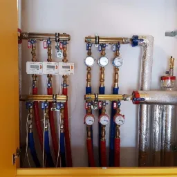 Rozdzielacz instalacji centralnego ogrzewania i wodnej z licznikami, rurami miedzianymi i plastikowymi, zabudowany w żółtej szafce, widoczne zawory i izolacja termiczna.