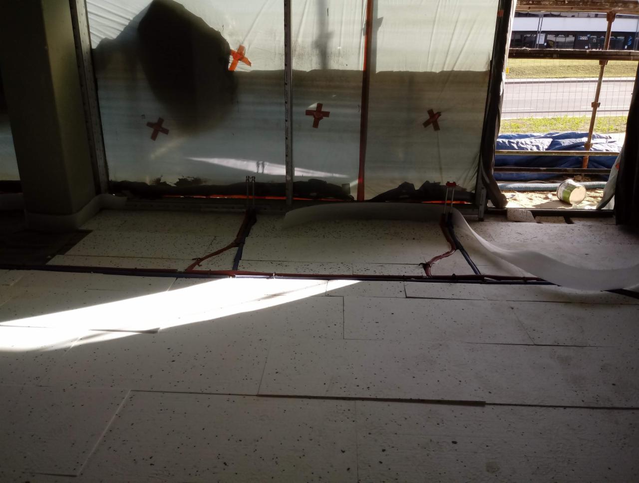 Podłoga w trakcie instalacji ogrzewania podłogowego, widoczne rury układane na białych płytach izolacyjnych, zabezpieczone folią okna i ściany, w tle widok na zewnątrz.