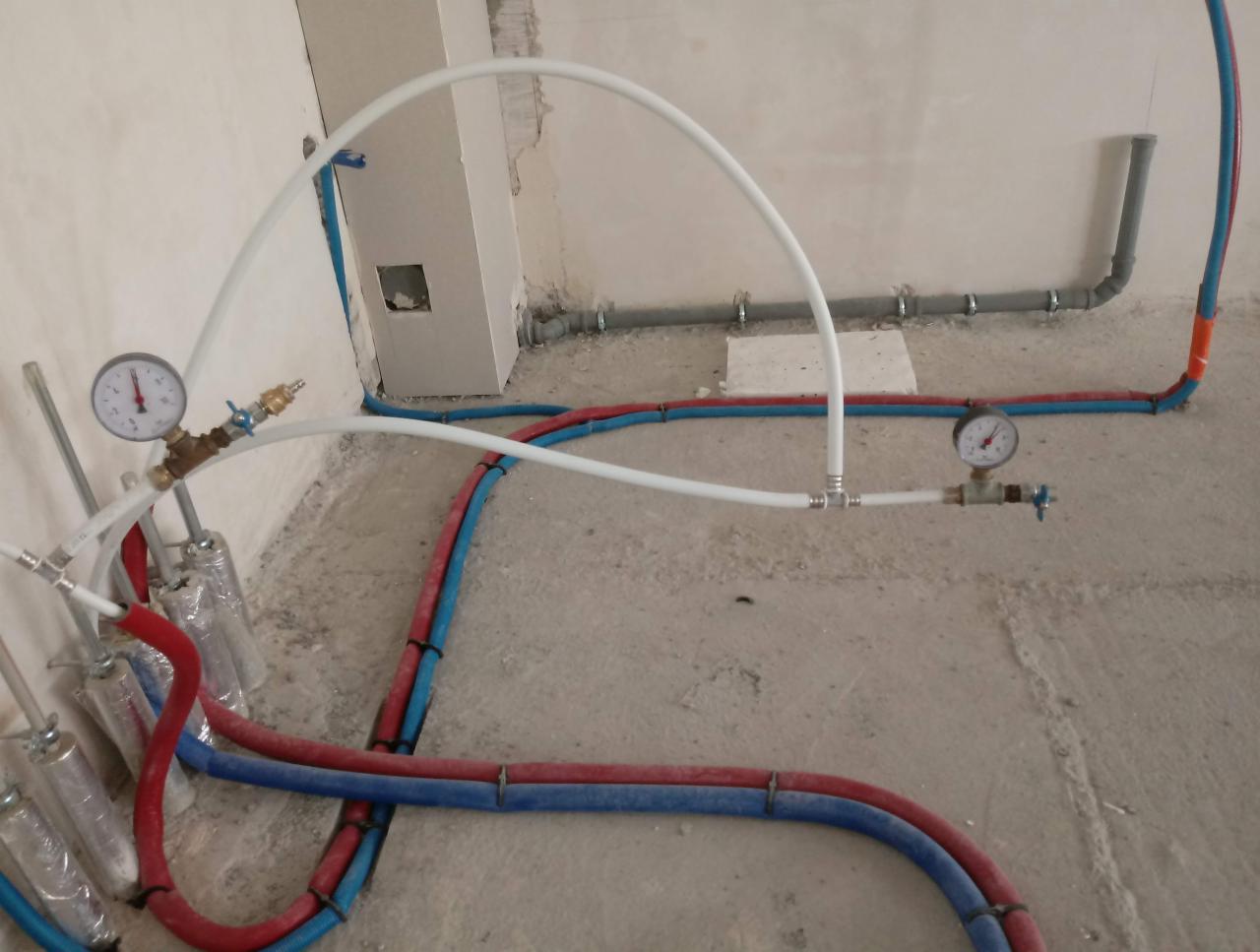 Wnętrze w trakcie prac hydraulicznych: widoczne rury w kolorach czerwonym i niebieskim, białe rury z manometrami, surowy beton na podłodze i ściany w trakcie wykończenia.