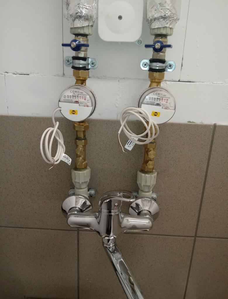 Bateria umywalkowa z dwoma wodomierzami na tle szaro-beżowej ściany z płytek, widoczne rury i zawory doprowadzające wodę.