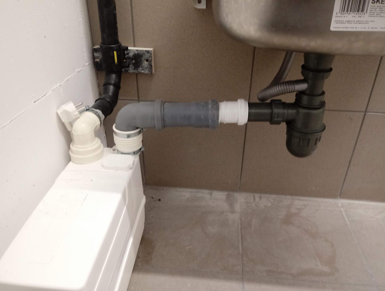 Instalacja odpływowa umywalki z zamontowaną pompą do ścieków szarych, widoczne rury PCV, syfon i biała obudowa pompy, podłączenie do kanalizacji w tle, płytki ceramiczne na ścianie i podłodze.