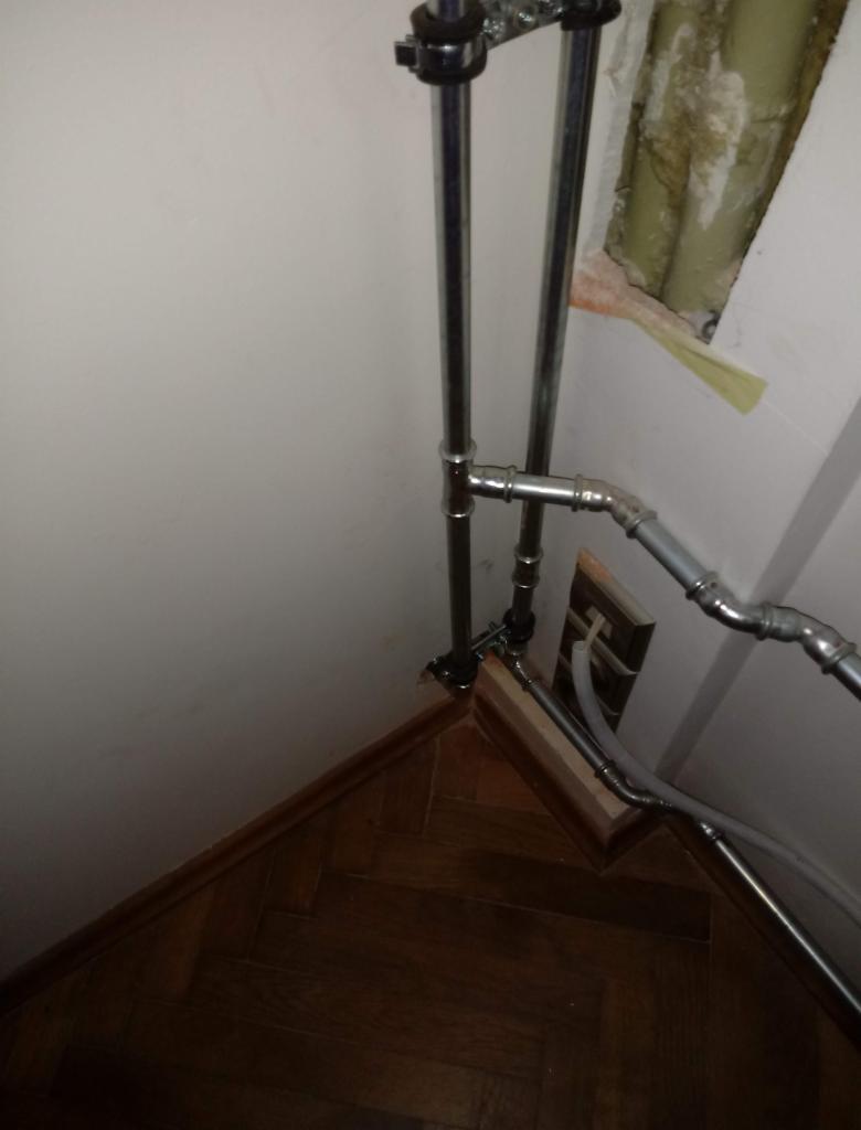 Metalowe rury instalacyjne, widoczne połączenia i mocowania, fragment drewnianej podłogi parkietowej, ściana z widocznym uszkodzeniem tynku.