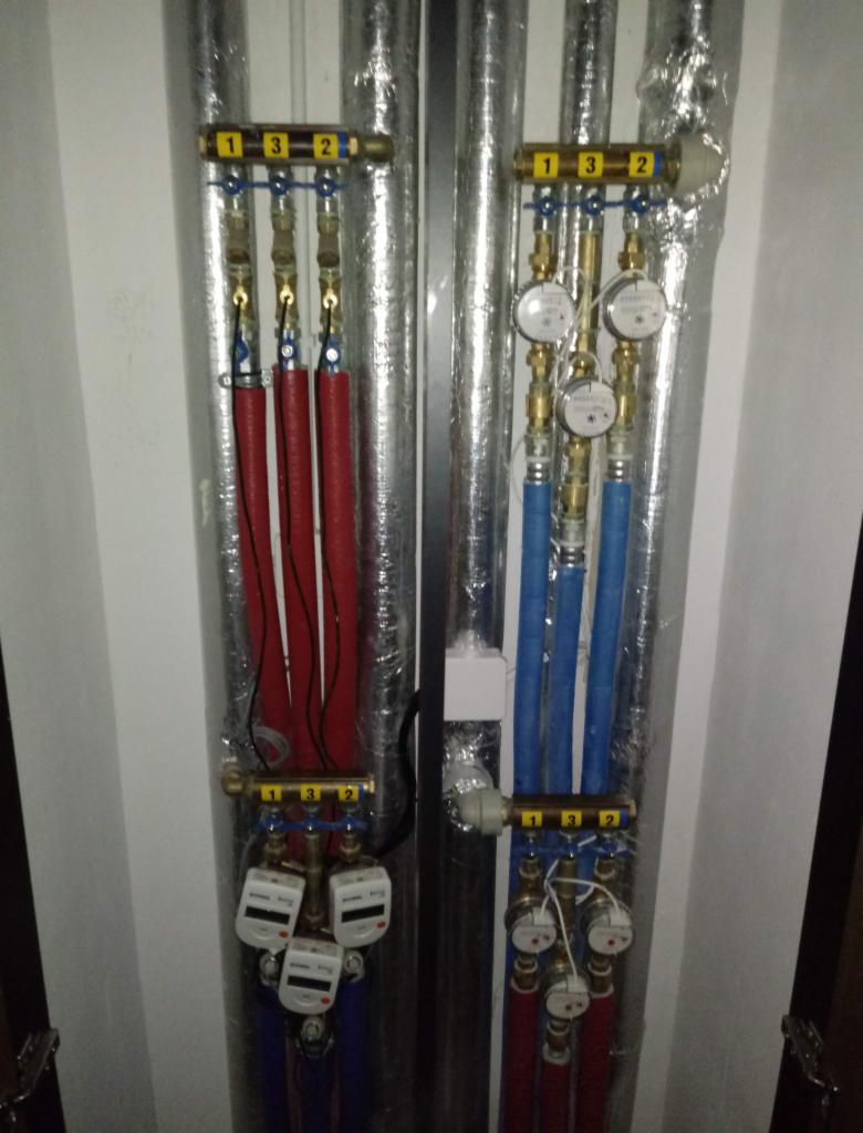 Rozdzielacz instalacji centralnego ogrzewania z rurami w izolacji, manometrami i licznikami zużycia, widoczne rury czerwone i niebieskie.